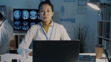 Beyaz önlüklü genç Asyalı kadın portresi karanlık ofiste dizüstü bilgisayarla masada oturuyor.