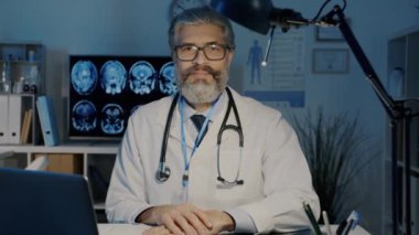 Yavaş çekimde, üniformalı, gülümseyen ve akşam ofisteki kameraya bakan dost canlısı erkek doktorun portresi.