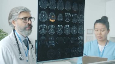 Nörolog MRI beyin taramalarını analiz ediyor ve modern klinikteki Asyalı hemşireyle konuşuyor.