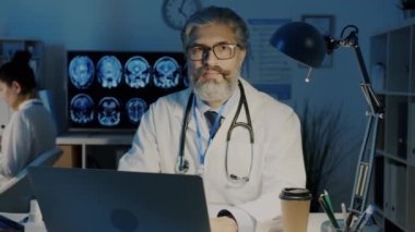 Tecrübeli erkek doktorun portresi masada bilgisayarla oturuyor ve geceleri hastanede kameraya bakıyor.