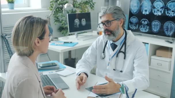 Doświadczony lekarz omawiający rezonans magnetyczny z pacjentką rozmawiającą w pokoju szpitalnym — Wideo stockowe