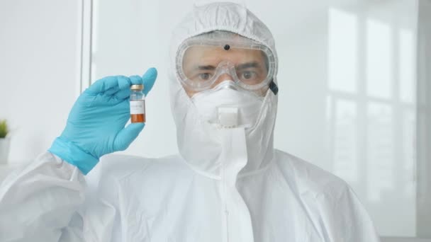 Retrato del médico varón en traje médico protector de pie en la unidad de aislamiento covid-19 que sostiene la vacuna mirando a la cámara — Vídeos de Stock
