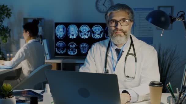 Slow motion portret van ervaren arts in witte jurk zittend aan bureau met laptop in donker kantoor — Stockvideo