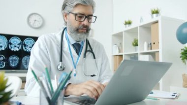 Hastanede tek başına çalışan, dizüstü bilgisayar kullanan beyaz önlüklü bir adamın portresi.