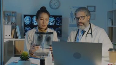 Asyalı kadın ve beyaz erkek doktorlar ciğerlerinin röntgen görüntüsünü analiz ediyor ve geceleri hastanede çalışıyor.