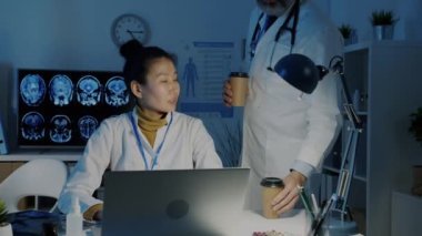 Yorgun Asyalı kadın doktor erkek meslektaşından kahve alıyor sonra da akşamları dizüstü bilgisayarla çalışıyor.