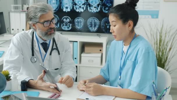 Медсестра в форме смотрит на экран планшета и разговаривает с врачом в белом халате в современной клинике — стоковое видео