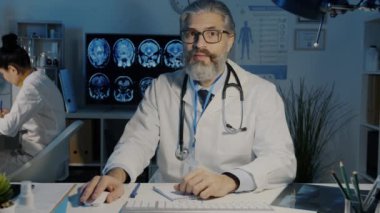 Deneyimli doktor konuşuyor ve karanlık ofiste internet danışmanlığı sırasında kameraya bakıyor.