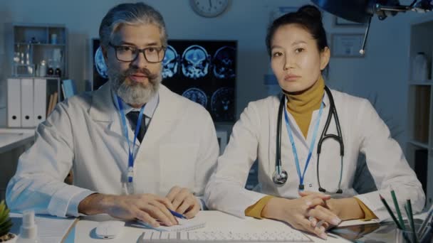 Mężczyzna i kobieta lekarze mówiący i gestykulujący patrząc w kamerę podczas wirtualnego spotkania z pacjentem w ciemnej klinice w nocy — Wideo stockowe