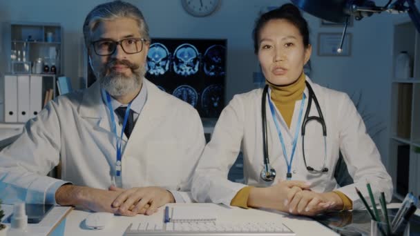 Азиатка и кавказский мужчина разговаривают и смотрят в камеру во время медицинского вебинара в темном офисе — стоковое видео