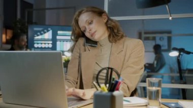 Genç bayan çalışan iş yerinde dizüstü bilgisayar kullanıyor ve cep telefonuyla konuşuyor.