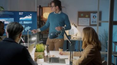 Karanlık ofisteki bir grup meslektaşına sunum yapan genç bir çalışan dijital ekranı işaret ediyor ve konuşuyor.