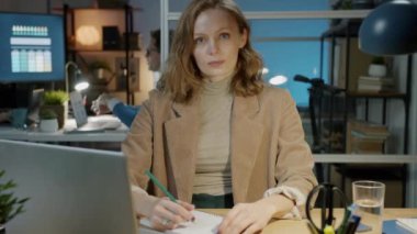 Ciddi bir iş kadınının kameraya bakıp karanlık yaratıcı ofiste masasında oturması.