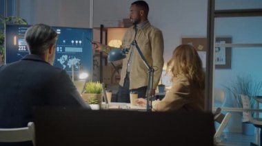 Karanlık ofiste sunum sırasında çeşitli meslektaşlarına konuşan Afrikalı Amerikalı bir çalışan.