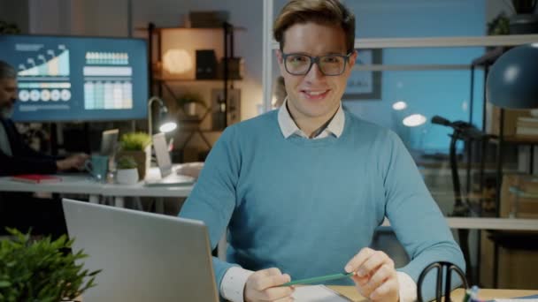Langzame beweging van vrolijke kantoormedewerker zittend achter computerbureau in donker kantoor glimlachend — Stockvideo