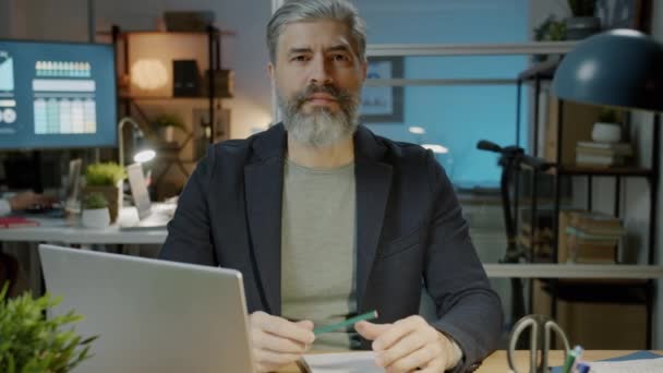 Portret van een serieuze ondernemer in formele kleding zittend aan een bureau in donker kantoor — Stockvideo