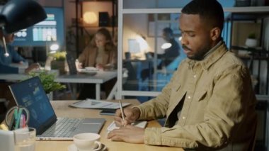 Afro-Amerikalı iş adamı dizüstü bilgisayara bakıyor not alıyor ve geceleri ofiste çalışırken kahve içiyor.