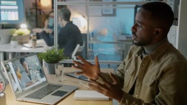 Genç işadamı kablosuz kulaklık takıyor ve gece ofisindeki video görüşmesi sırasında iş arkadaşıyla konuşuyor.