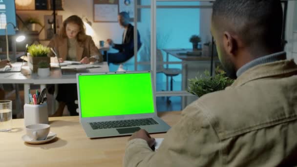Sobre o ombro tiro de empresário afro-americano olhando para laptop com tela verde chave chroma no escritório escuro — Vídeo de Stock