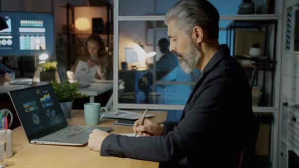 Vista lateral del hombre de negocios escribiendo mirando la pantalla del ordenador portátil haciendo investigación de marketing en la oficina oscura por la noche — Vídeo de stock