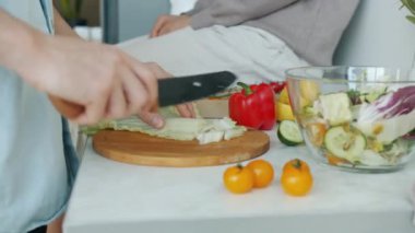 Kadın evde mutfak masasında otururken erkek eller salata için sebze kesiyor.