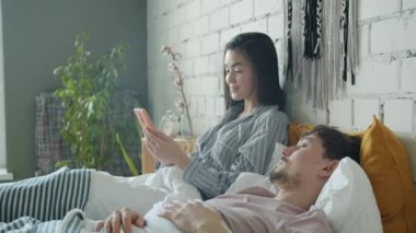 Akıllı telefon kullanan Asyalı kadın ekranda gülümserken melez ırk adamı evde uyuyor.
