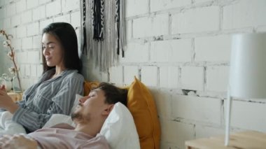 Mutlu Asyalı kadın akıllı telefonuyla eğlenirken erkek arkadaşı evde uyuyor.