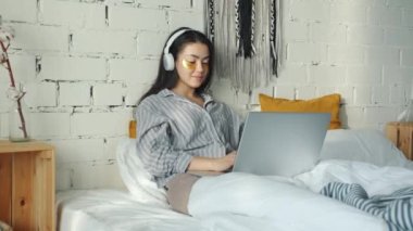 Göz bantlı Asyalı kadın kulaklıkla müzik dinliyor ve evde dizüstü bilgisayarla yazı yazıyor.