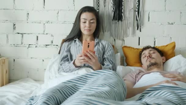 아시아의 젊은 여자가 남편 이 집에서 침대에서 잠을 자는 동안 스마트폰 문자를 사용하는 모습 — 비디오