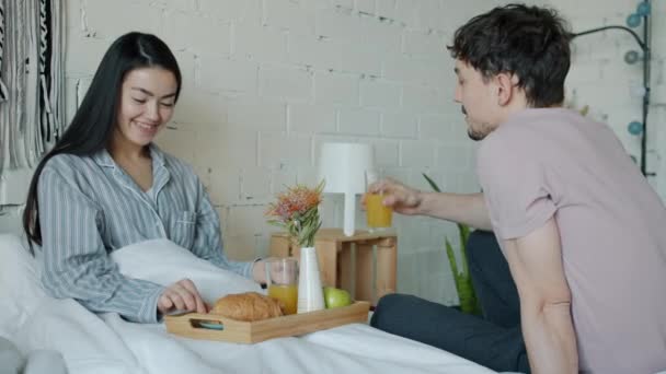 Asiática señora alimentación joven marido con croissant y beber jugo disfrutando de desayuno en cama — Vídeo de stock