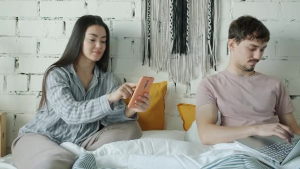 Lány és srác használ okostelefon és laptop pihentető ágyban otthon — Stock videók