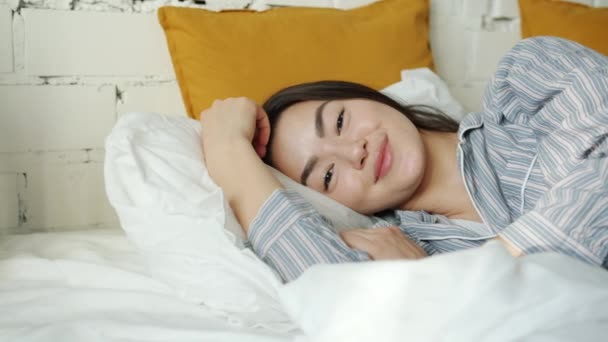 Retrato de la alegre señora asiática con pijamas acostada en la cama en casa sonriendo mirando a la cámara — Vídeo de stock