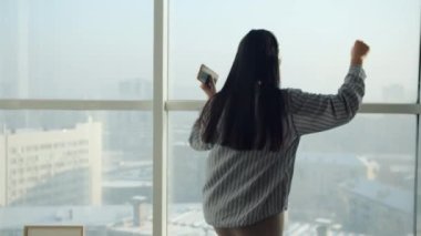 Kulaklık takan Asyalı bir kadının evde panoramik pencere önünde dans edip eğlenmesi.