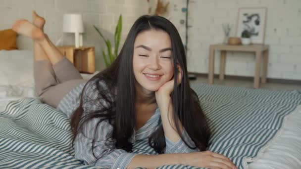 Retrato en cámara lenta de una atractiva mujer asiática acostada en la cama sonriendo mirando a la cámara en casa — Vídeos de Stock