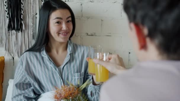 Девушка и парень звонят в стаканы с соком и едят круассан наслаждаясь завтраком в постели дома — стоковое видео