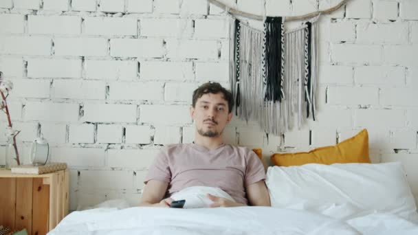 Портрет расового человека, смотрящего телевизор, меняющего каналы, чувствующего разочарование в постели дома — стоковое видео