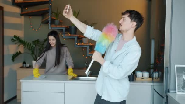 Jovem com espanador posando para câmera de smartphone se divertindo enquanto dona de casa limpeza cozinha no fundo — Vídeo de Stock