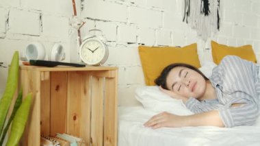 Asyalı bir kadın uyuyor sonra da evdeki yatak odasında çalar saati kapatıyor.