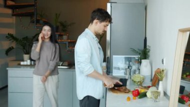 Genç adam yemek pişirirken, kadın mutfakta cep telefonuyla konuşuyor.