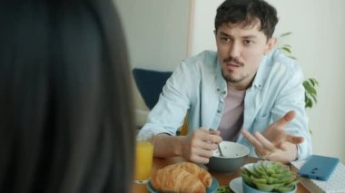 Mutlu genç adam kahvaltı yapıyor ve mutfakta oturan kadınla konuşuyor.