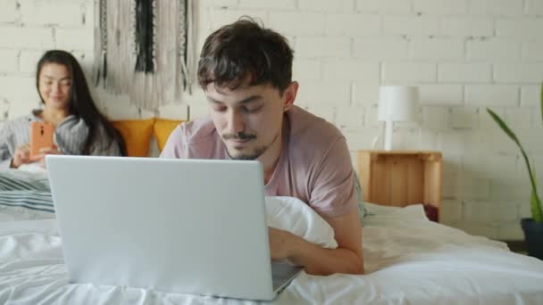 Marido trabajando con el ordenador portátil de escribir mientras que la esposa usando el teléfono inteligente en la cama en el apartamento — Vídeo de stock