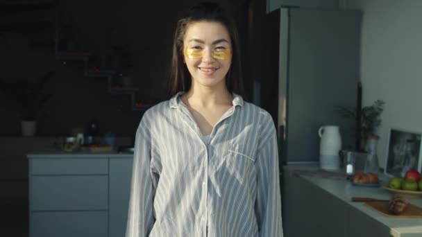 Retrato de la alegre chica asiática con pijamas y ojos parches sonriendo de pie en la cocina en casa — Vídeos de Stock