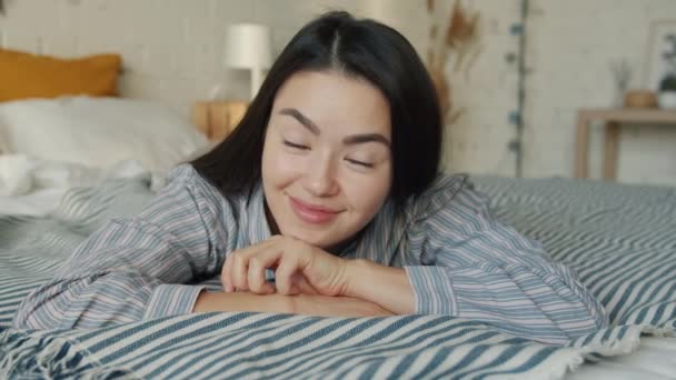 Retrato de una hermosa estudiante asiática acostada en la cama sonriendo y mirando a la cámara — Vídeo de stock