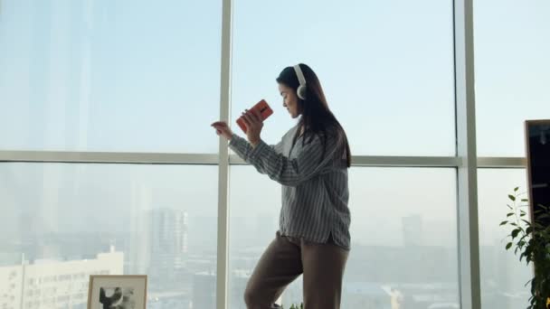 Наклон азиатки, танцующей дома в наушниках, держащей смартфон на фоне панорамного окна — стоковое видео