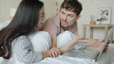Dizüstü bilgisayar kullanan erkek ve kadın evde yatakta başparmak göstererek içeriği tartışıyor.