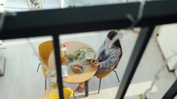 Genç bayanın kahvaltı edişinin ve evdeki mutfak masasında akıllı telefon mesajı kullanışının en üst görüntüsü. — Stok video