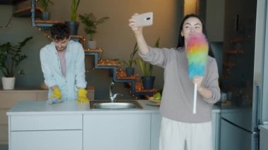 Erkek evi temizlerken ve gülümserken akıllı telefon kamerasına poz veren tozlu genç bir kadın.