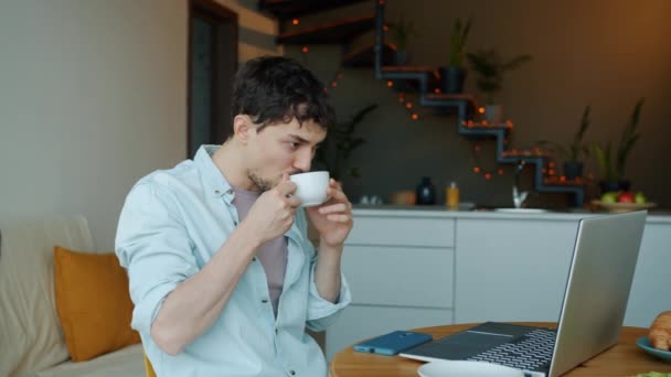 Pensive τηλεργασία πίνοντας καφέ και κοιτάζοντας οθόνη laptop στο τραπέζι της κουζίνας στο σπίτι — Αρχείο Βίντεο