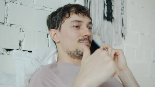 Portrait d'un gars heureux bavardant sur un téléphone portable souriant montrant un geste pouce levé au lit à la maison — Video