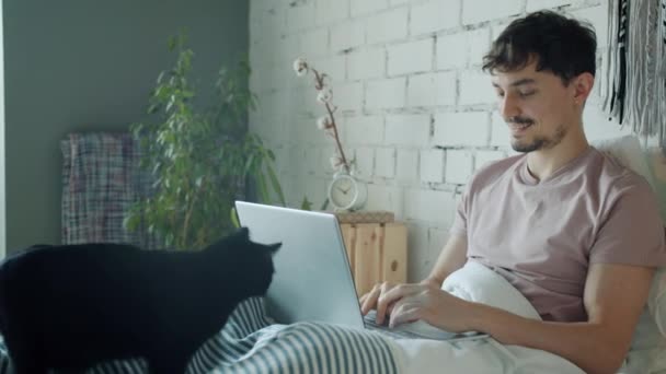 Genç adam dizüstü bilgisayar kullanıyor ve cep telefonuyla konuşuyor evde kediyi okşuyor. — Stok video